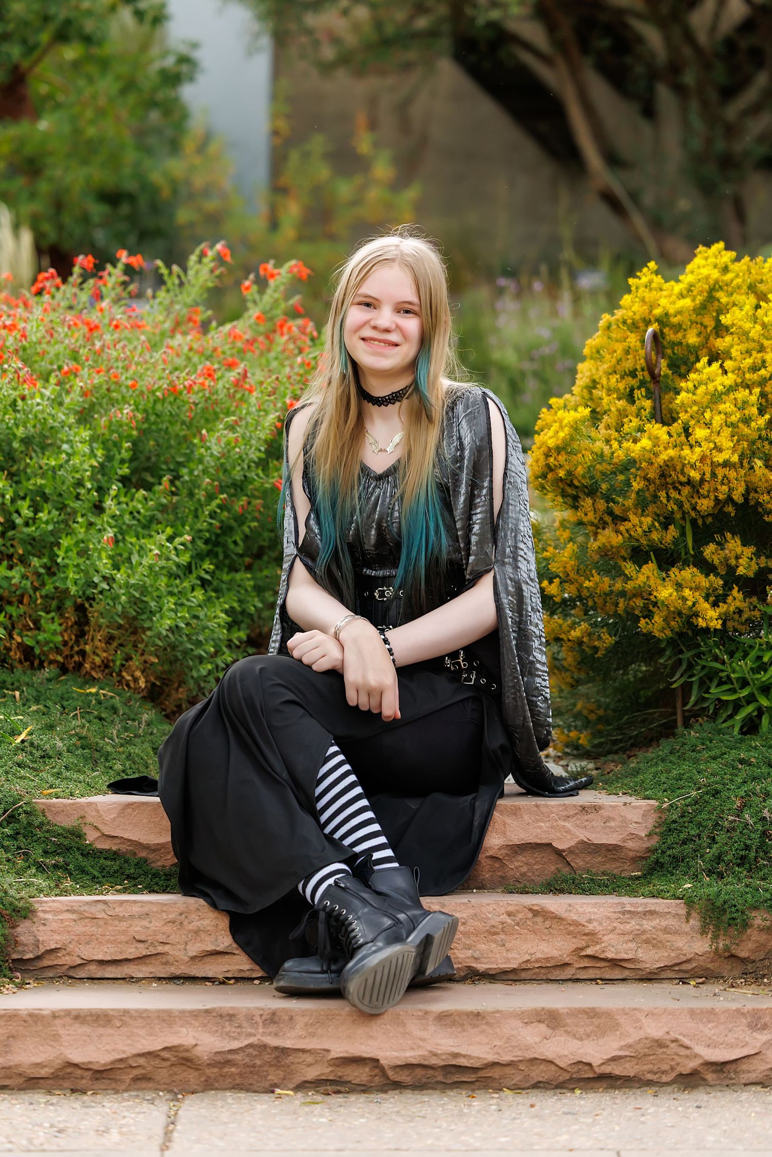 Young woman with blonde and teal hair sits on stone steps in a garden, wearing black outfit and striped socks.