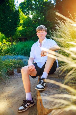 Outdoor portrait in sunlit garden with ornamental grasses showing someone seated on a log wearing white shirt and sneakers.