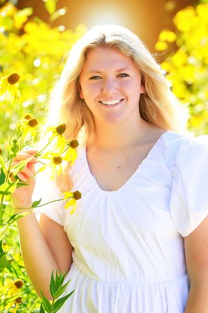 A bright outdoor portrait in a white dress surrounded by yellow flowers and sunlit foliage.
