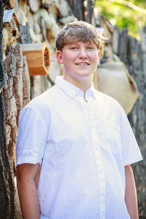 A person wearing a white button-down shirt stands outdoors among tree trunks on a sunny day.