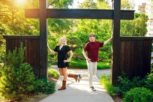 Two people jumping joyfully under a wooden garden archway surrounded by greenery on a sunny day.