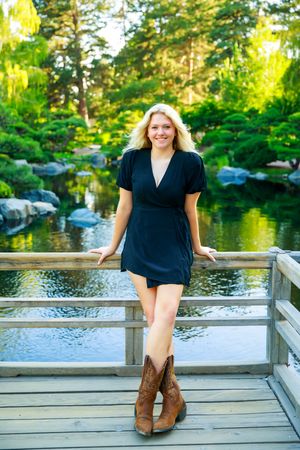 Person in navy wrap dress and brown cowboy boots poses on wooden bridge over tranquil Japanese garden pond.