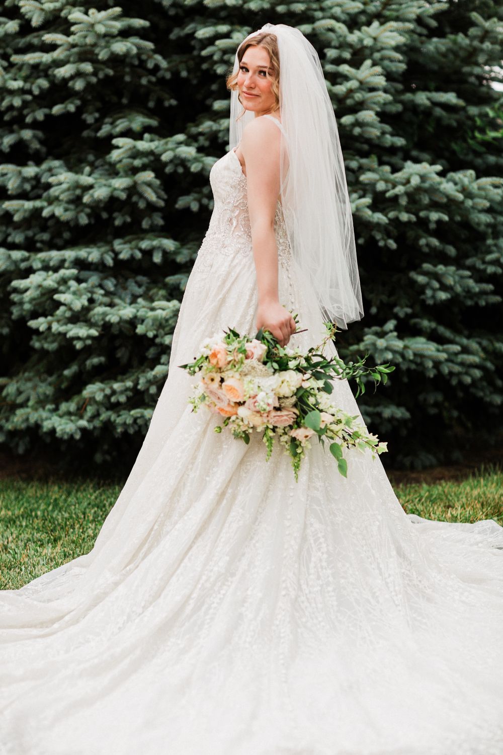 Bride in lace gown with long train poses with floral bouquet against evergreen backdrop.