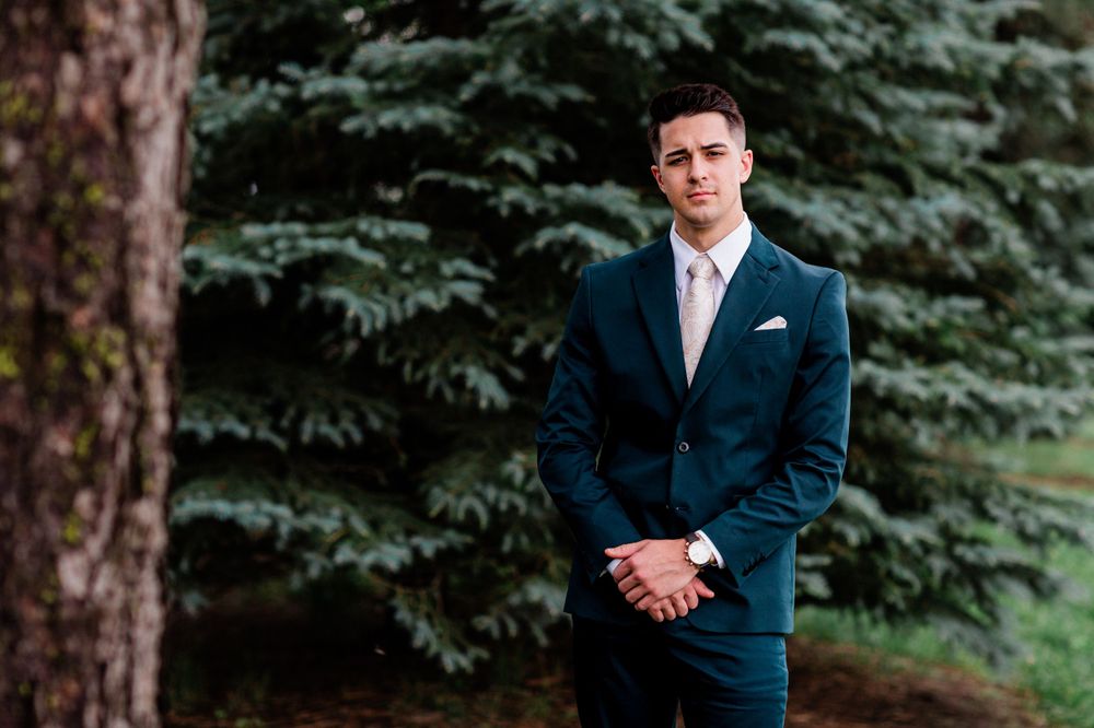 Groom in navy blue suit stands formally against evergreen trees.