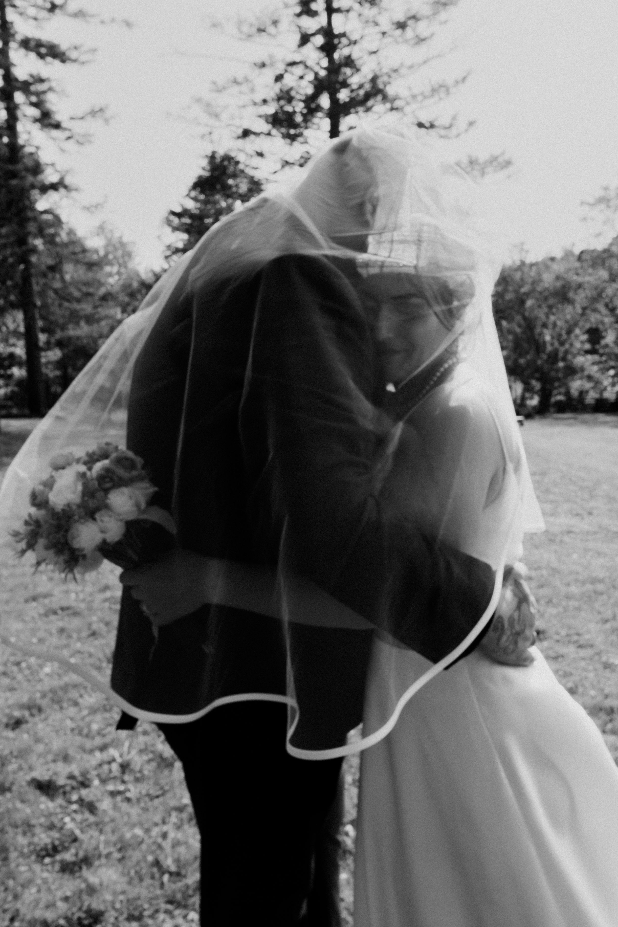 A romantic black and white portrait of newlyweds embracing under a flowing veil.