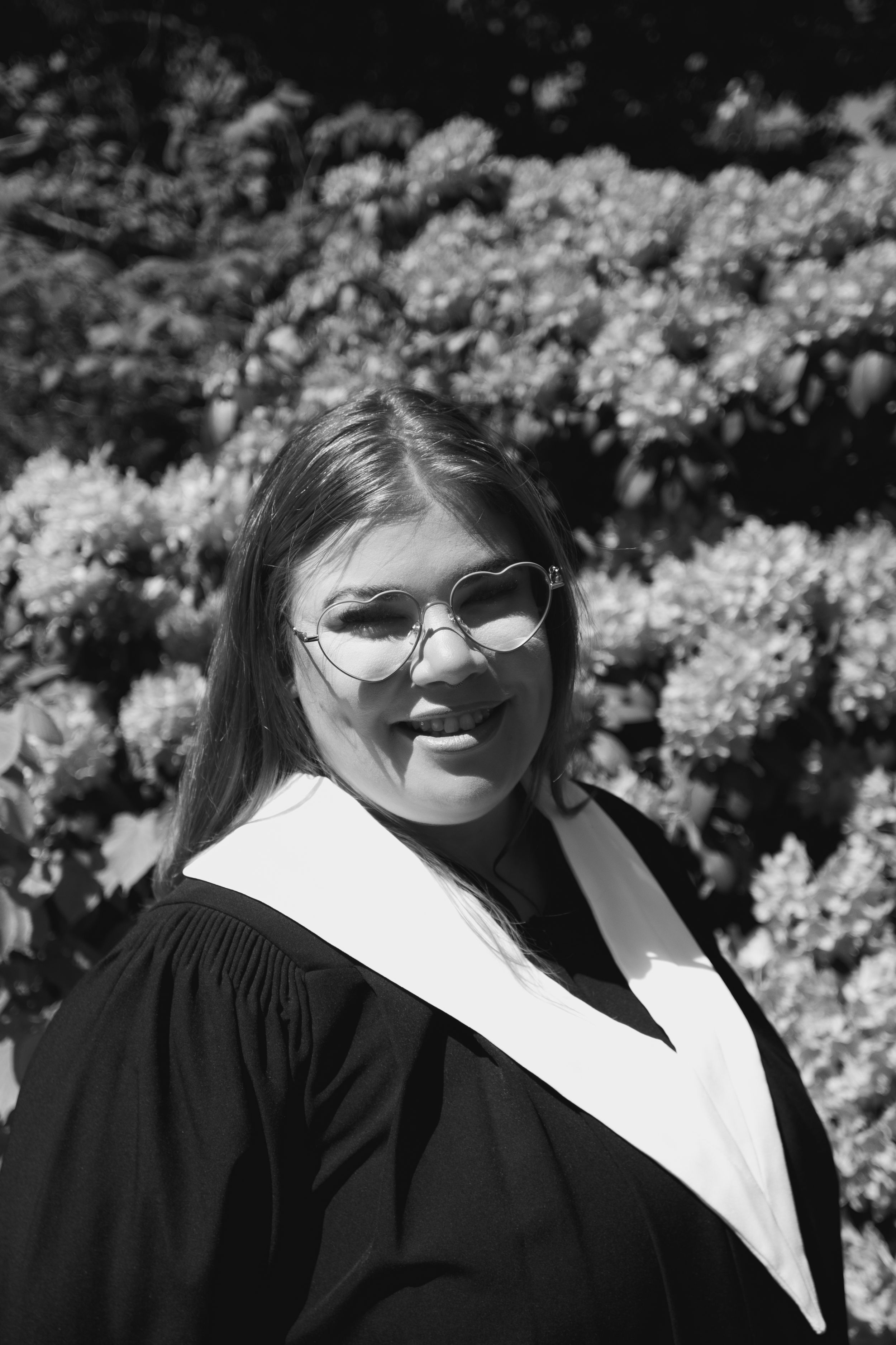 A graduate in cap and gown smiling in front of blooming flowers.