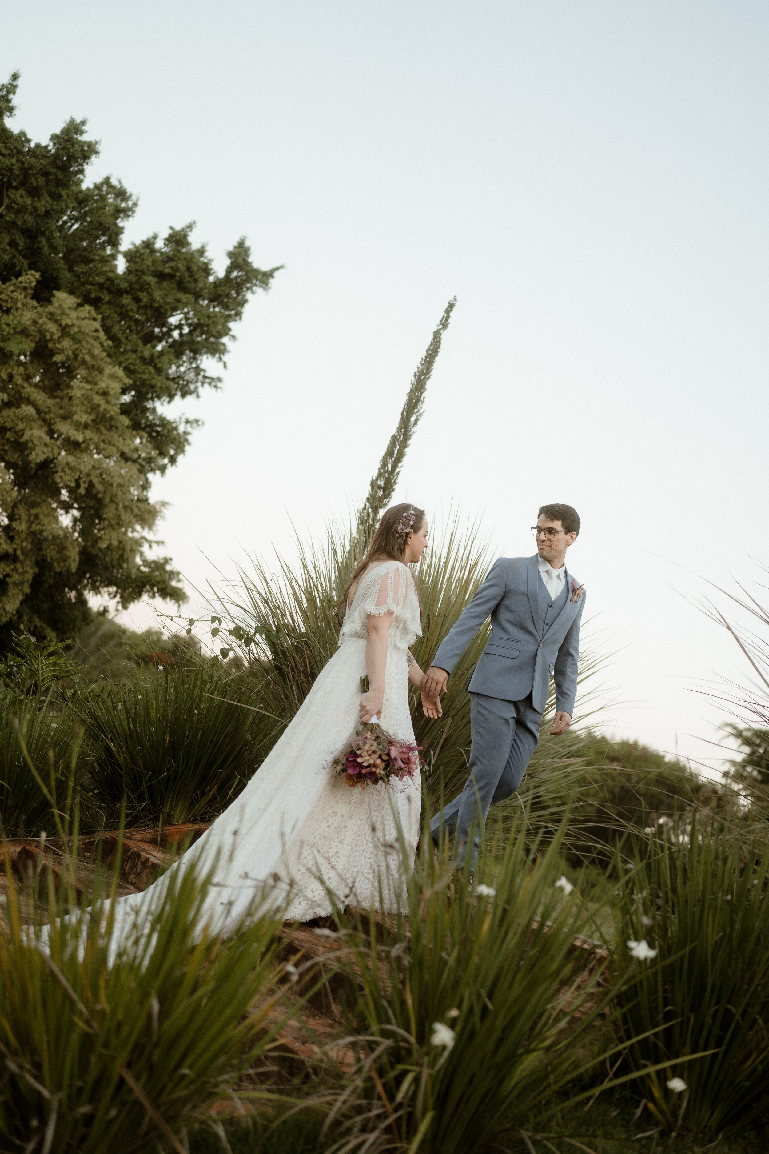 Newlyweds in formal attire pose together among tall grass and trees at sunset.