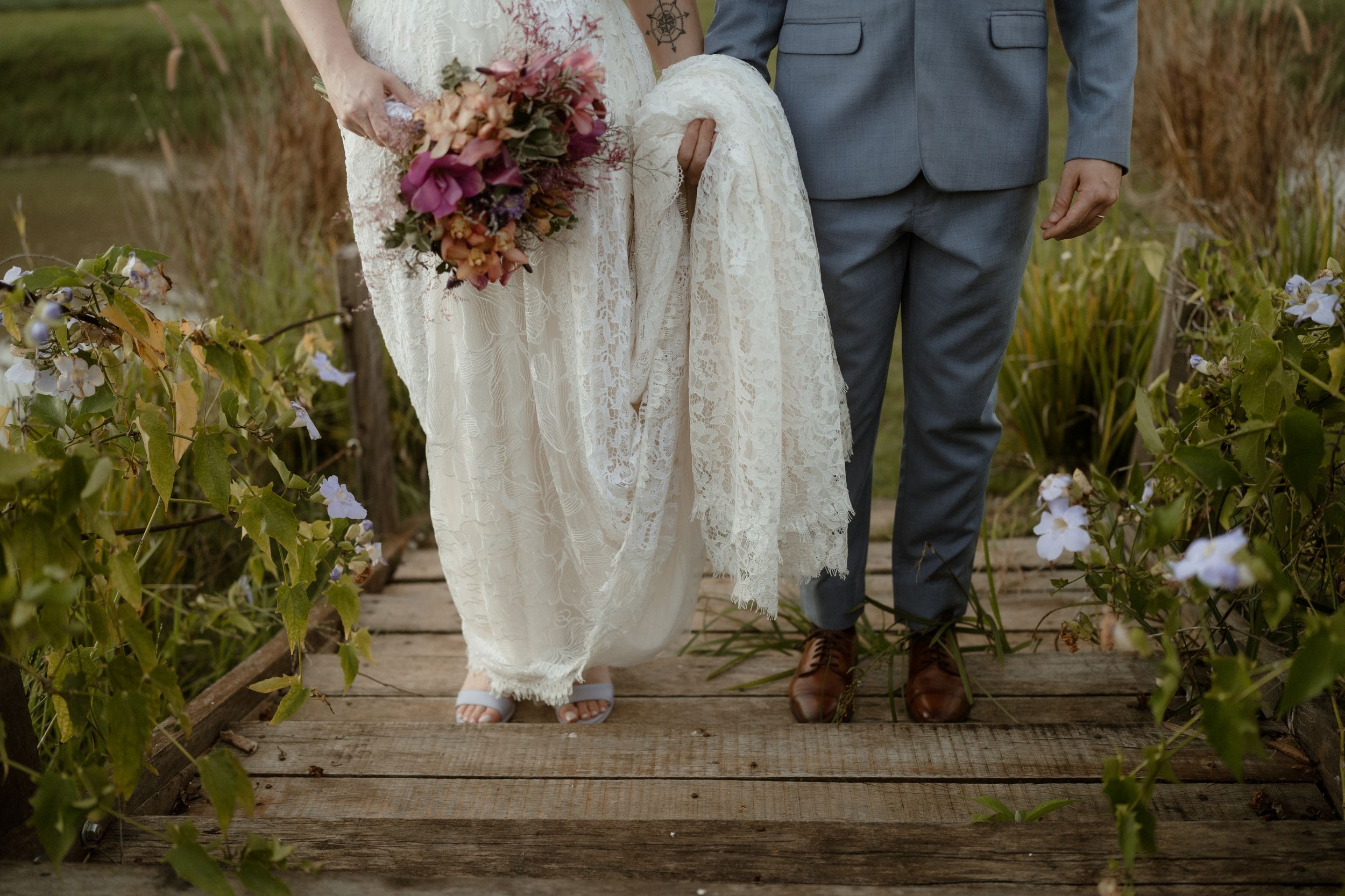 A bride in a lace dress and groom in gray suit stand on a wooden walkway holding a pink and purple bouquet.
