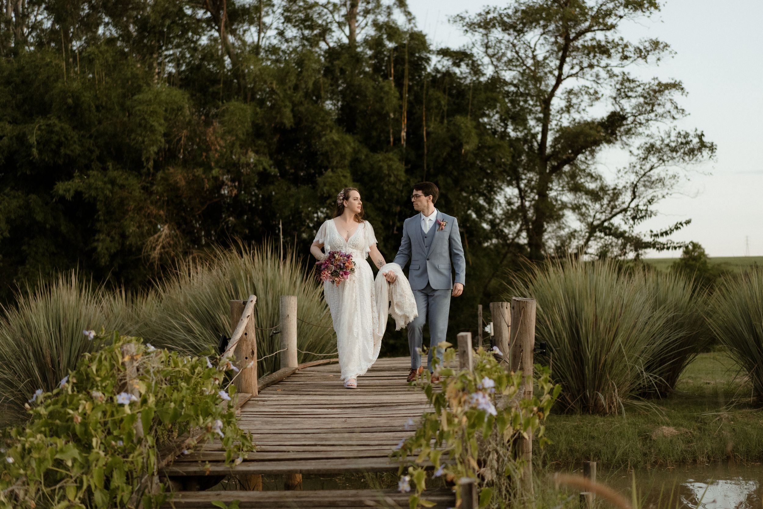 A couple walks together on a wooden boardwalk surrounded by coastal vegetation.