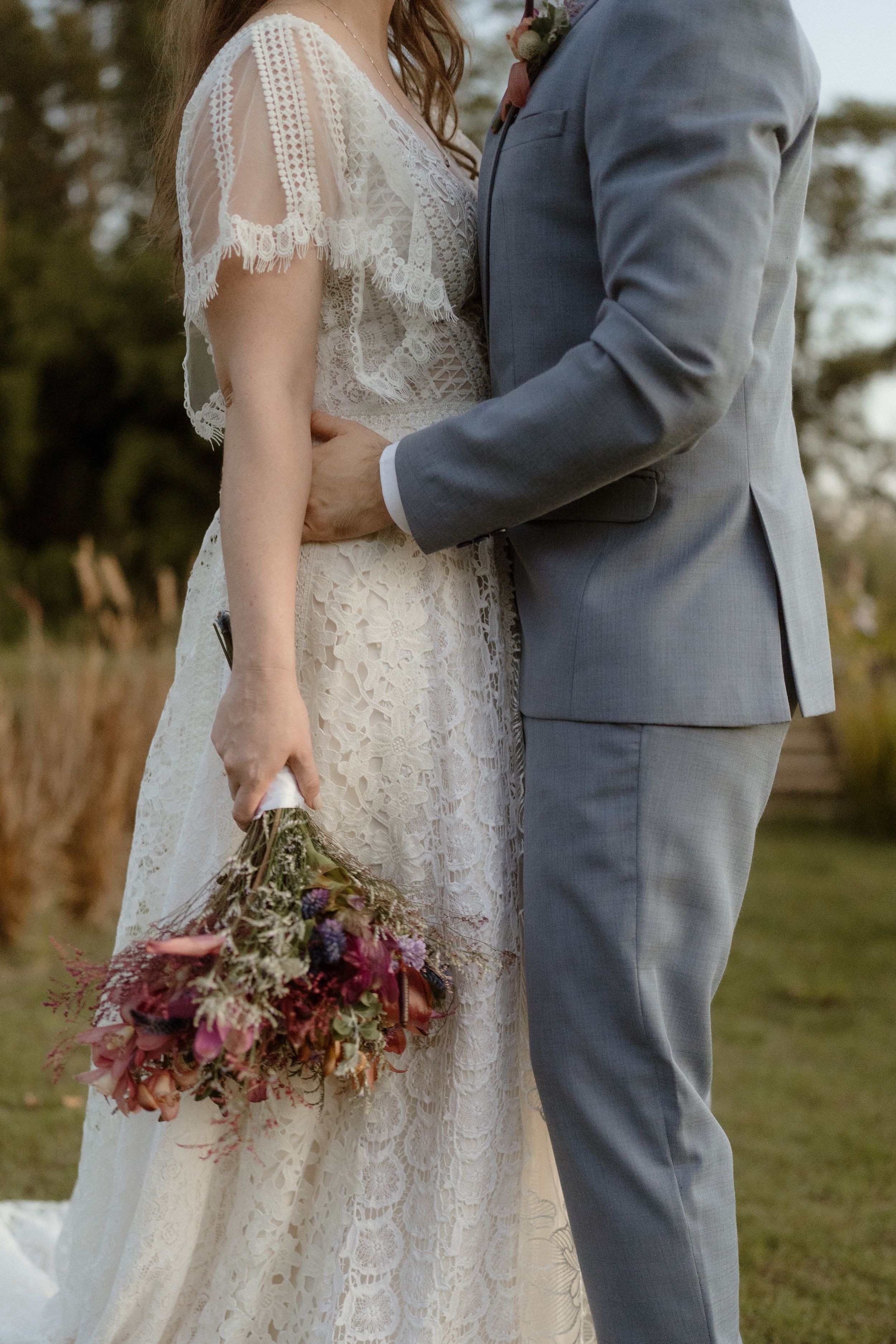 Close up of bride in lace dress holding dried flower bouquet next to groom in gray suit.