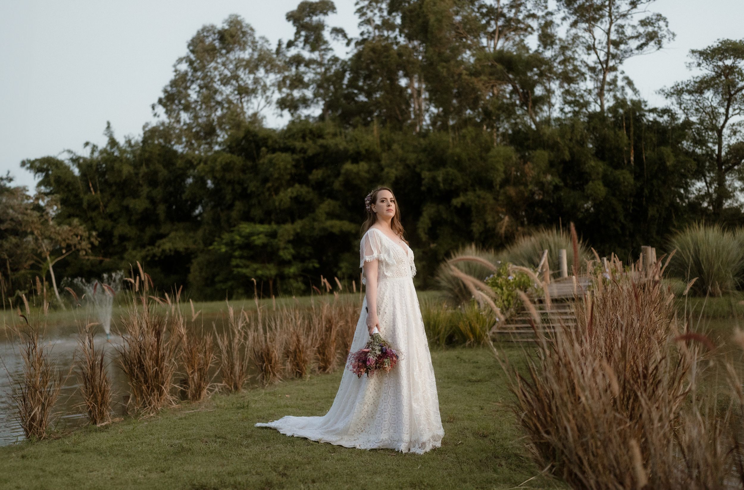Bride in flowing lace dress poses in garden setting with dried floral bouquet.
