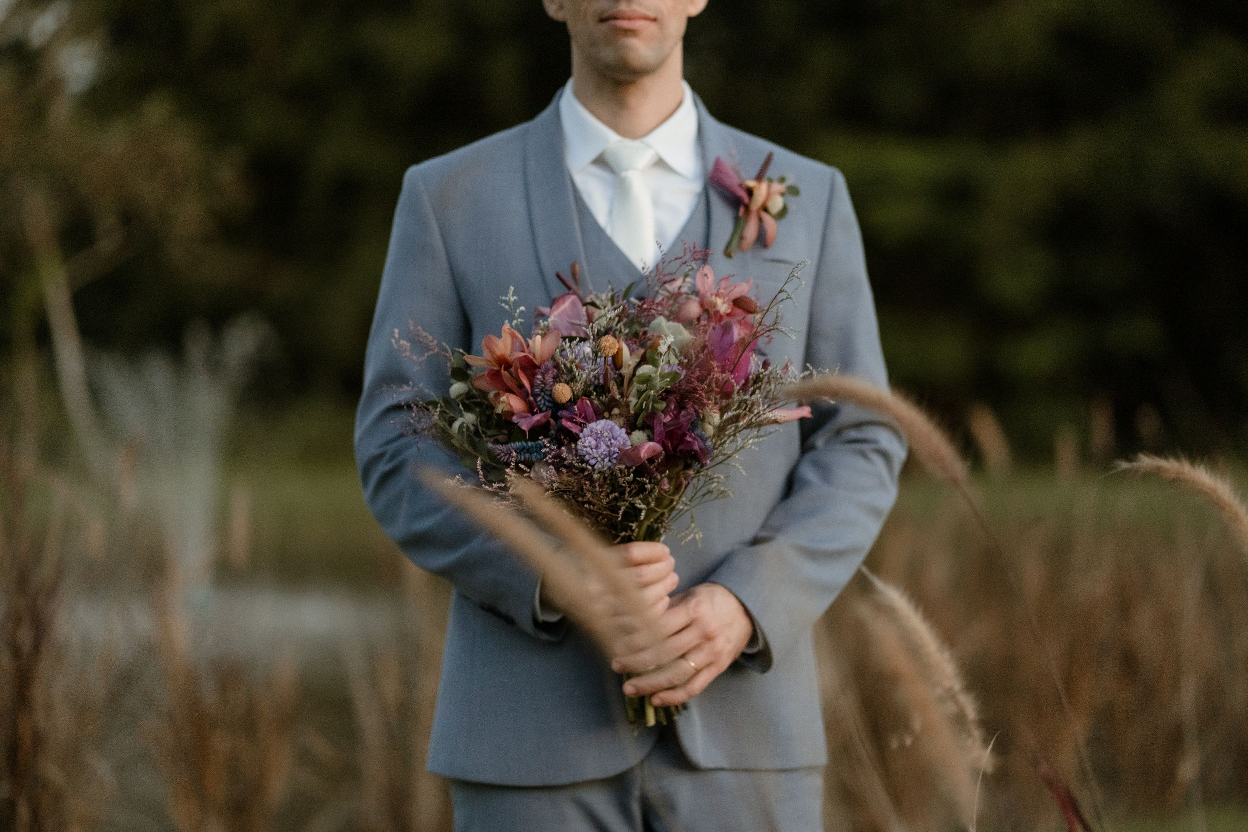 Groom in light blue suit holding pink and purple dried flower bouquet against natural backdrop.
