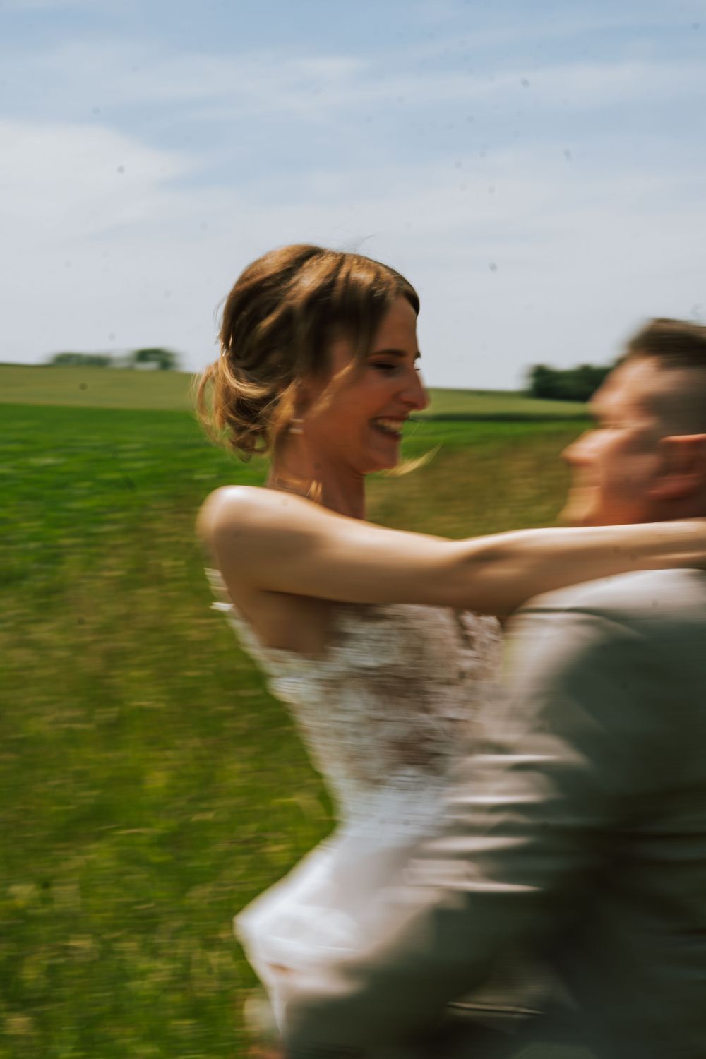 Couple in wedding attire spinning joyfully in a green field.
