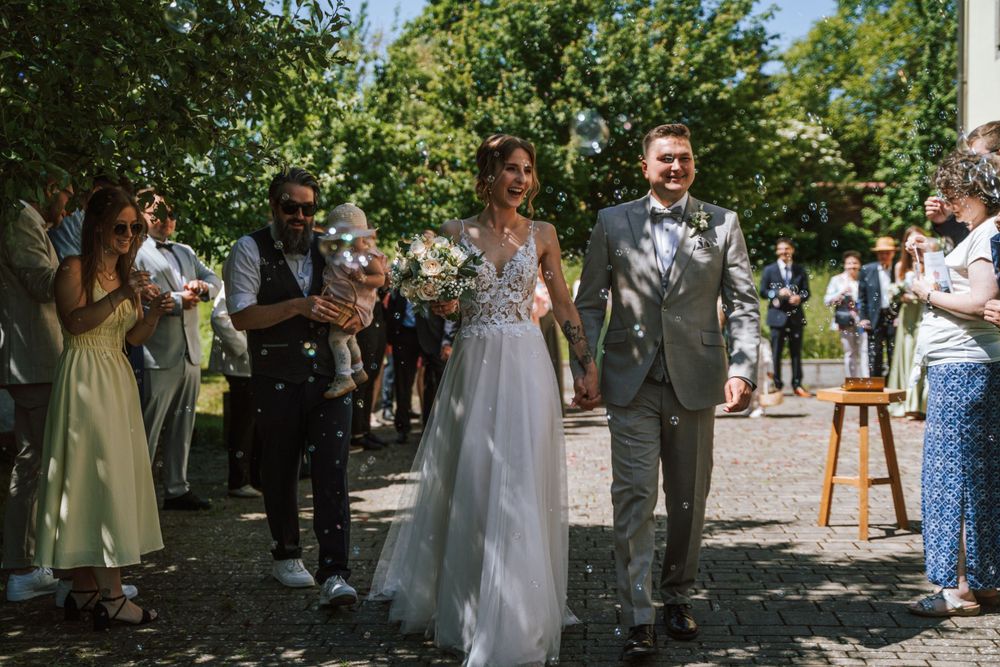 Wedding party walks outdoors among guests on sunny garden pathway.