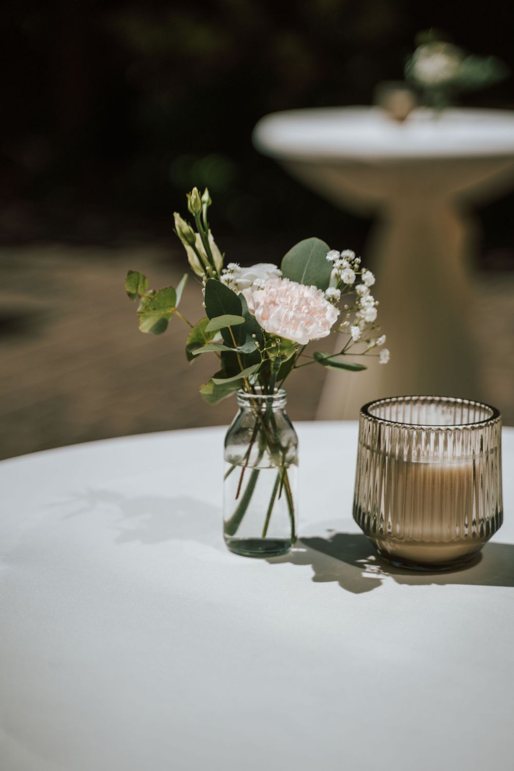 A simple wedding centerpiece with pink flowers in a glass vase and candle holder on white tablecloth.
