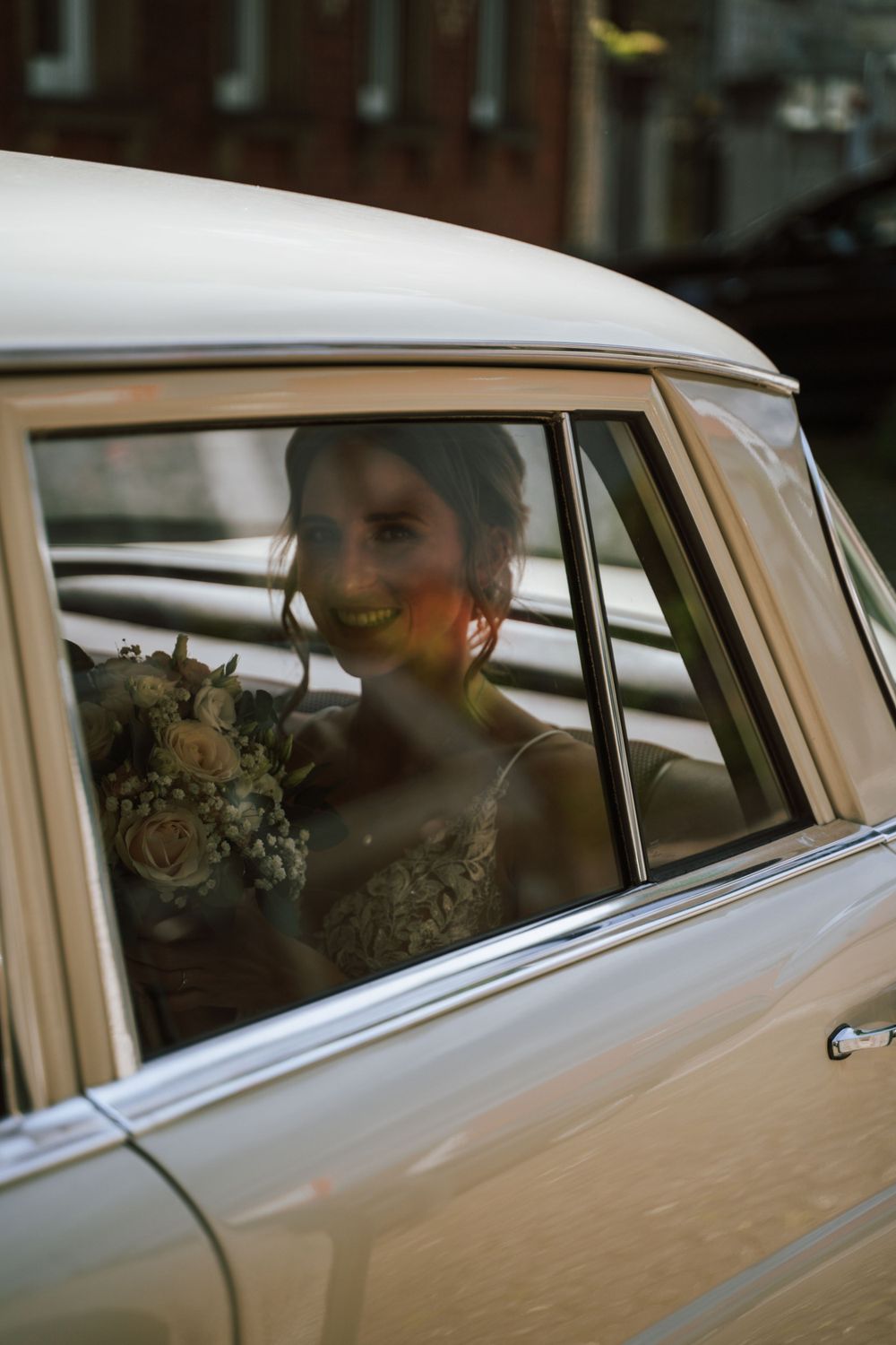 A vintage car window frames the elegant wedding dress and bouquet.