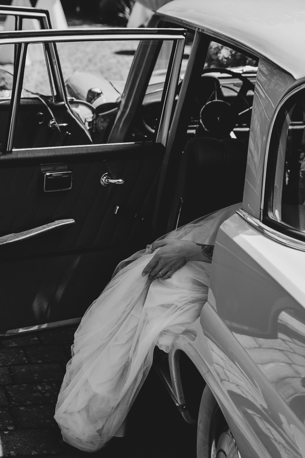 A black and white photo of a wedding dress flowing out of a classic car door.