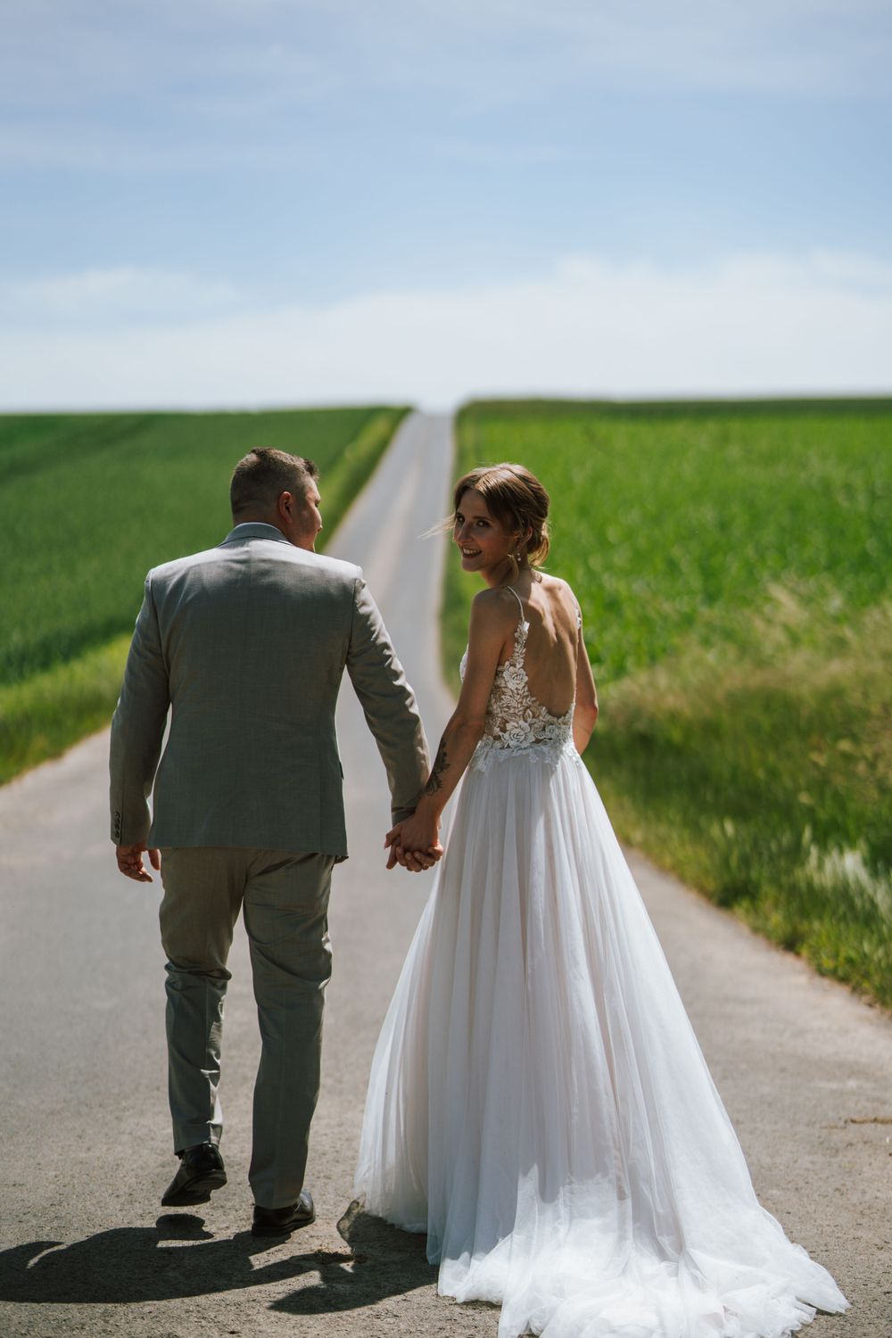 A couple walks hand in hand down a rural road surrounded by green fields.