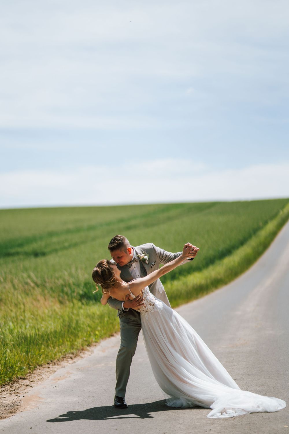 A romantic wedding couple dips dramatically on a country road surrounded by green fields.