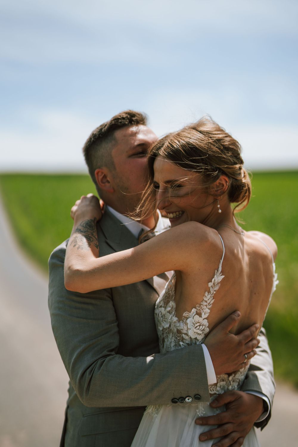 Romantic embrace between newlyweds on a rural road with green fields in background.