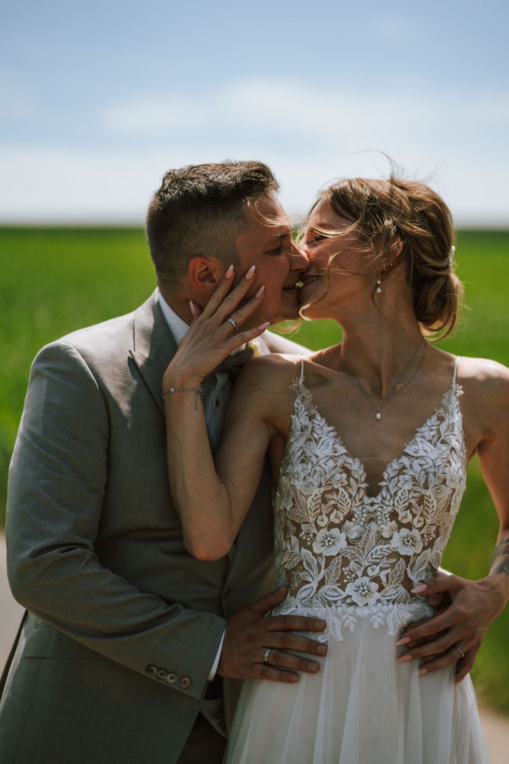 A romantic wedding kiss between newlyweds in a lush green field under blue skies.