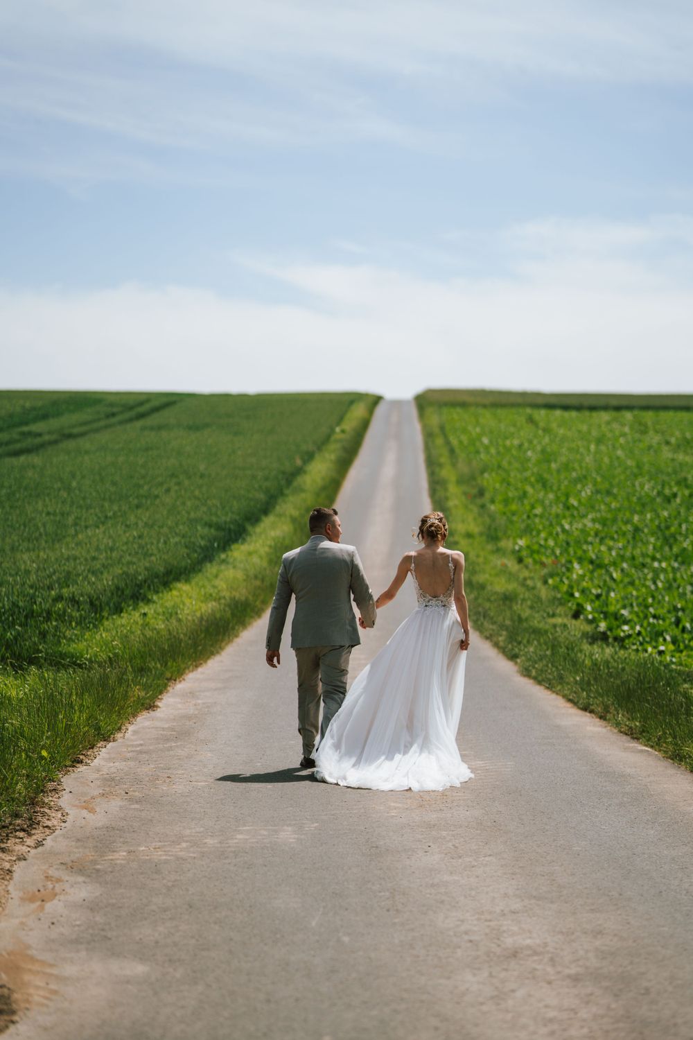 Newlyweds walking hand in hand down a country road between green fields.