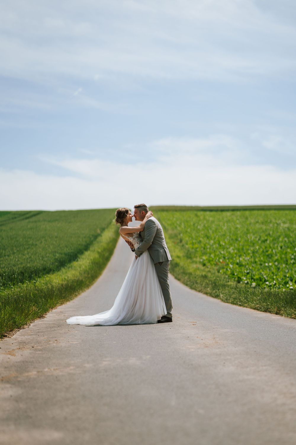 A romantic embrace between newlyweds on a rural road surrounded by green farmland.