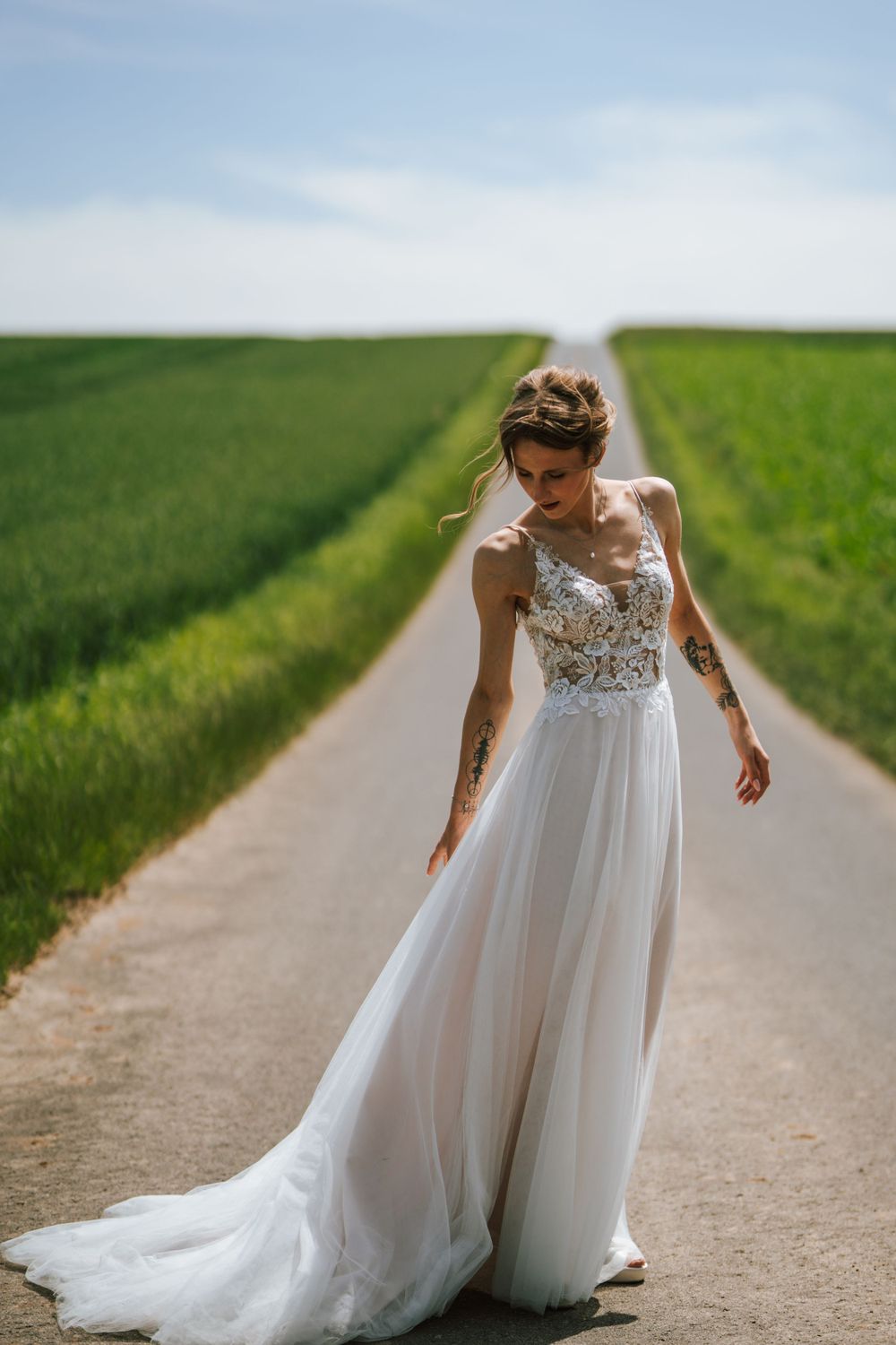 A bride in a flowing lace wedding gown stands on a country road between green fields.
