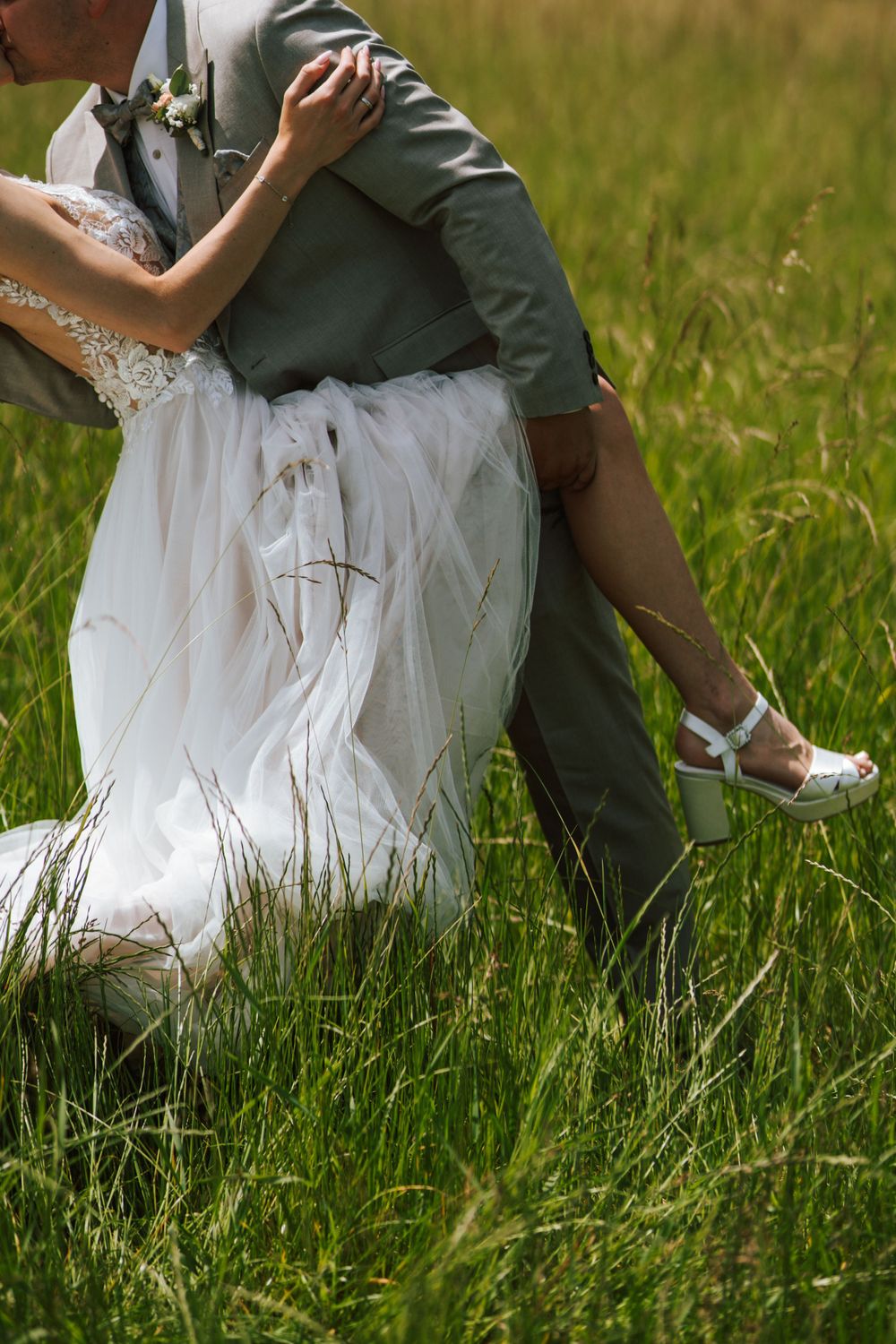 Intimate moment between newlyweds sitting in tall grass, showing wedding dress and grey suit details.