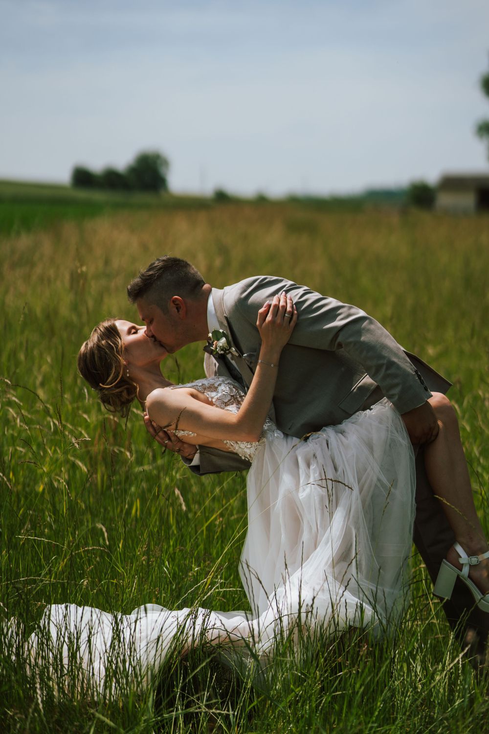 Romantic dip kiss between newlyweds in a summer field with golden light.
