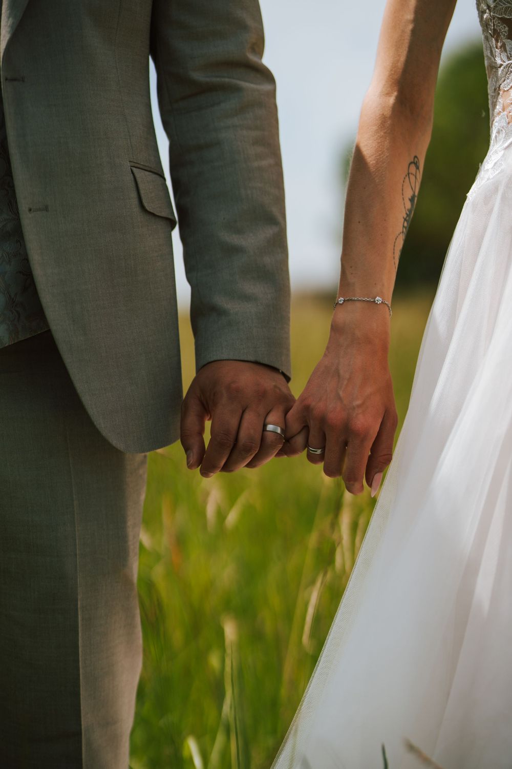 Hands touching white wedding dress fabric against grey suit in outdoor setting.