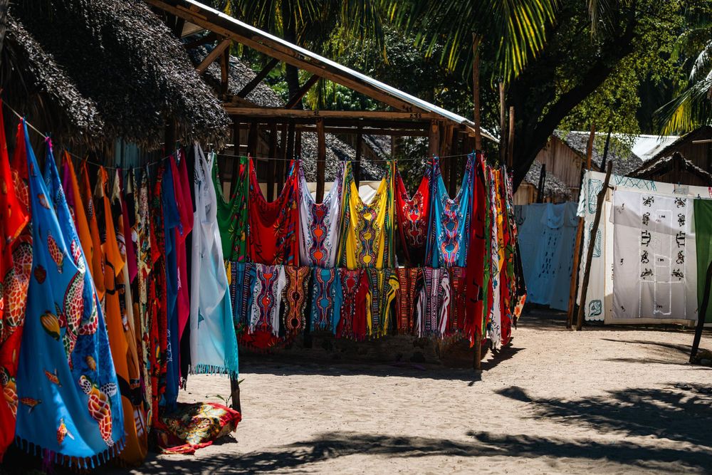 Colorful sarongs and beach wear hanging at an outdoor market stall under a thatched roof.