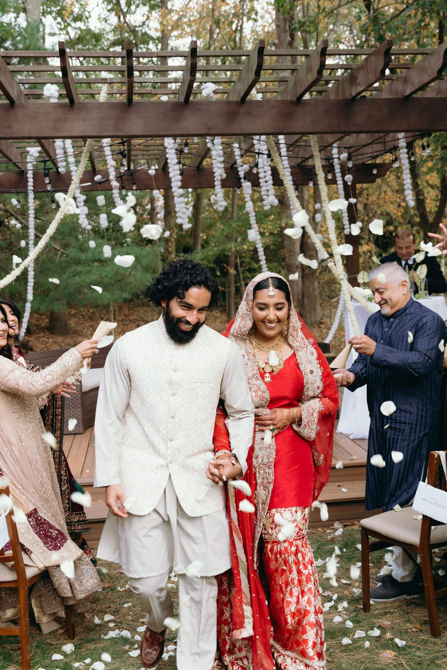 Indian bride in red lehenga and groom in white sherwani walk down the aisle as guests toss flower petals.