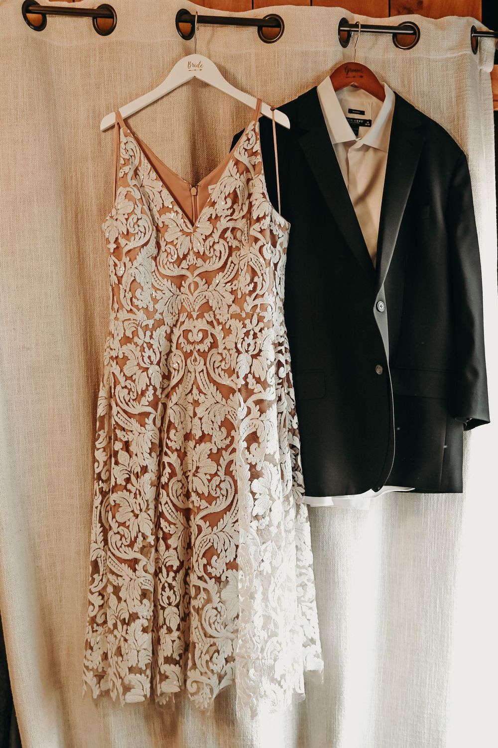 A delicate white lace wedding dress hangs next to a dark suit jacket on wooden hangers.