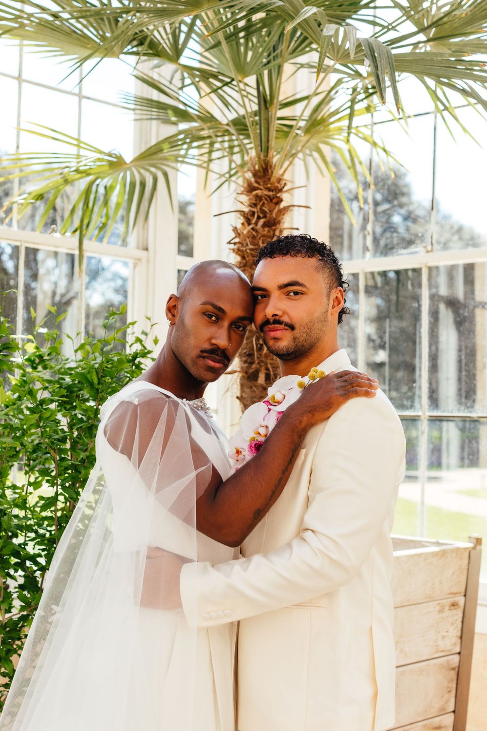 Two people in white wedding attire embrace under a palm tree in a sunlit conservatory.