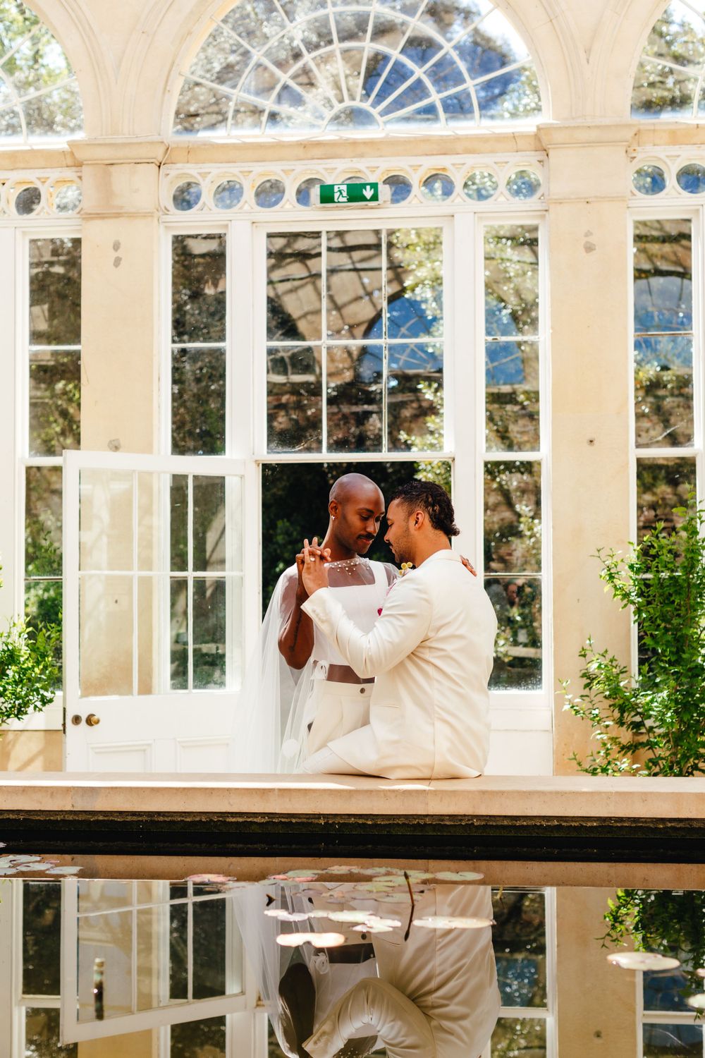 Couple in elegant wedding attire share a romantic moment in a glass-windowed conservatory.