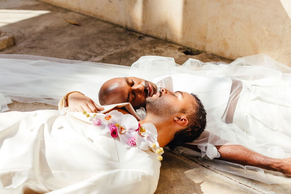 Two people in white wedding attire lay together peacefully in dappled sunlight.