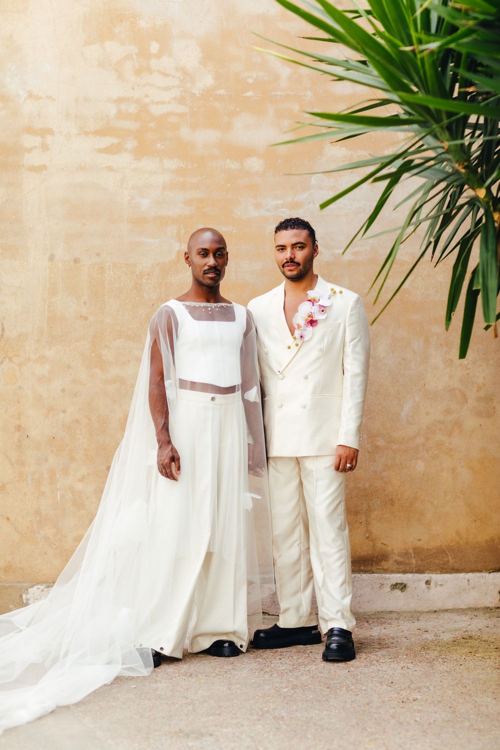 Wedding portrait of couple in coordinating white outfits against beige wall with palm leaves.