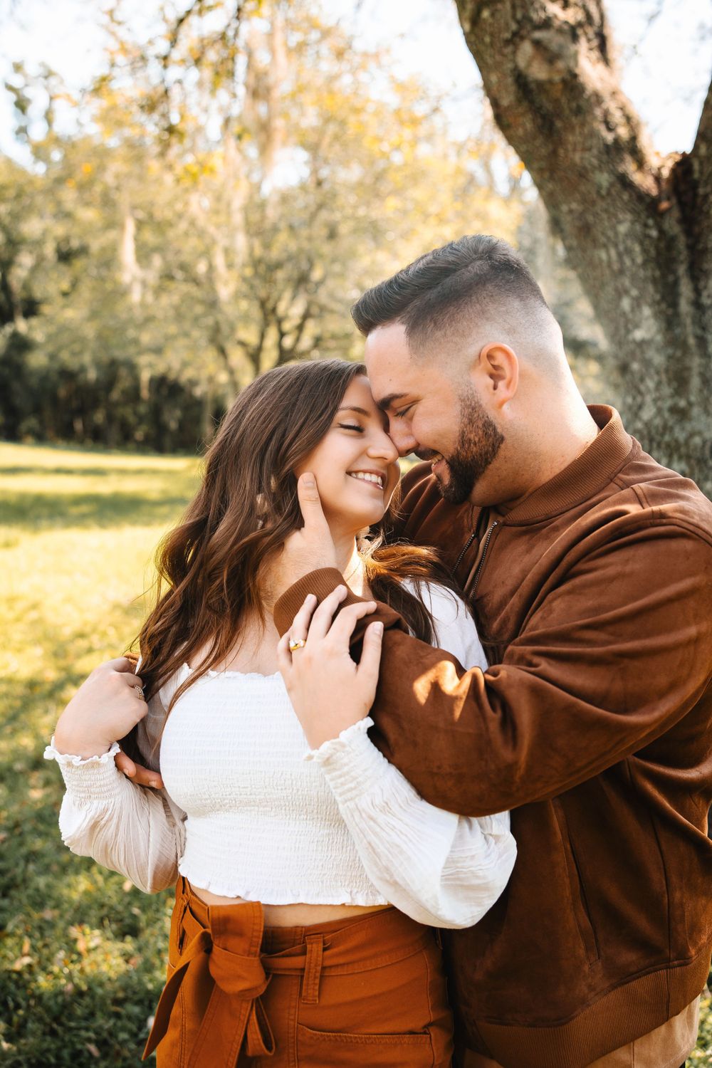 Couple sharing an intimate moment outdoors, woman smiling as man gently touches her face near oak tree.