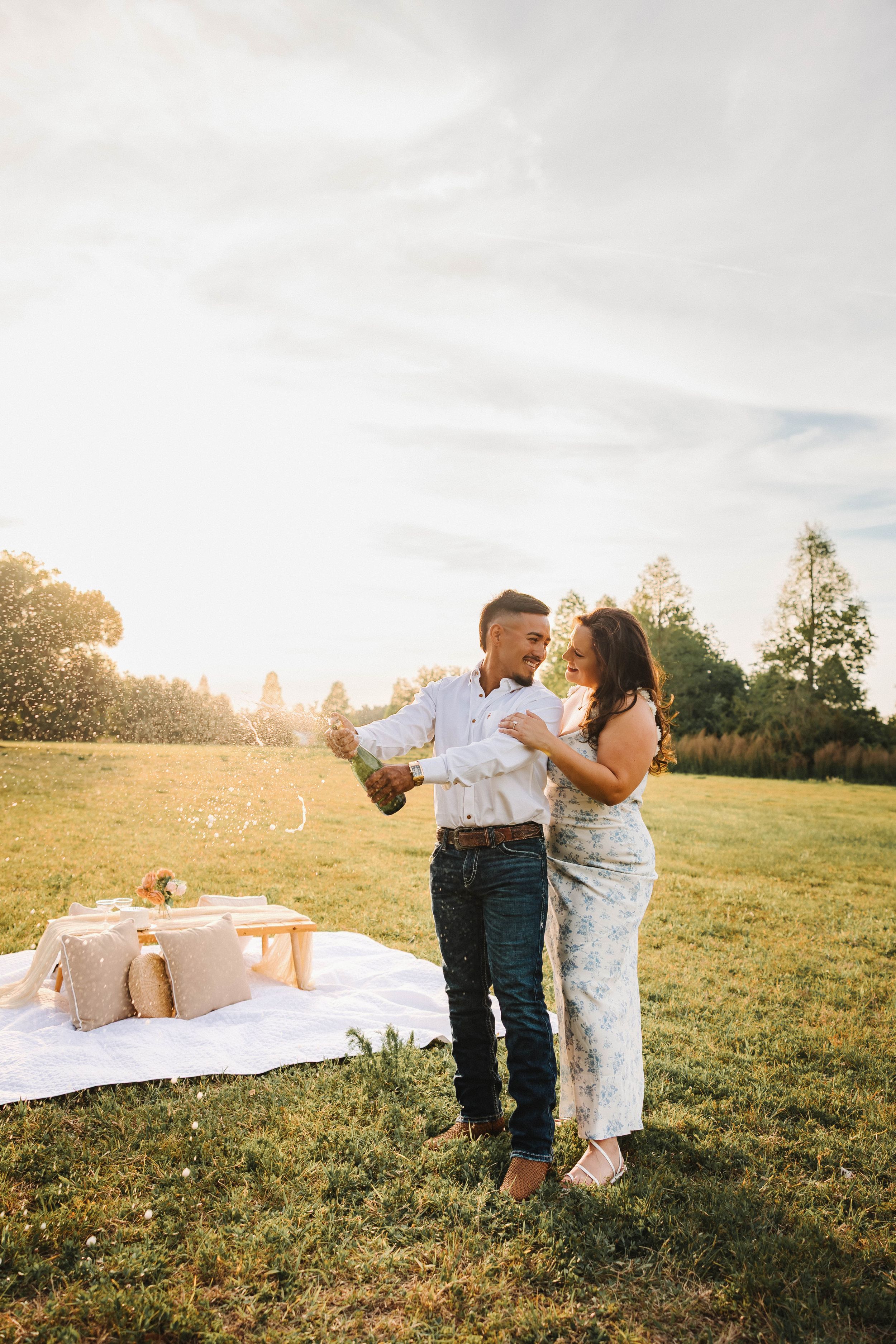 Couple celebrating outdoors at golden hour, popping champagne on a picnic blanket in a sunny green field.