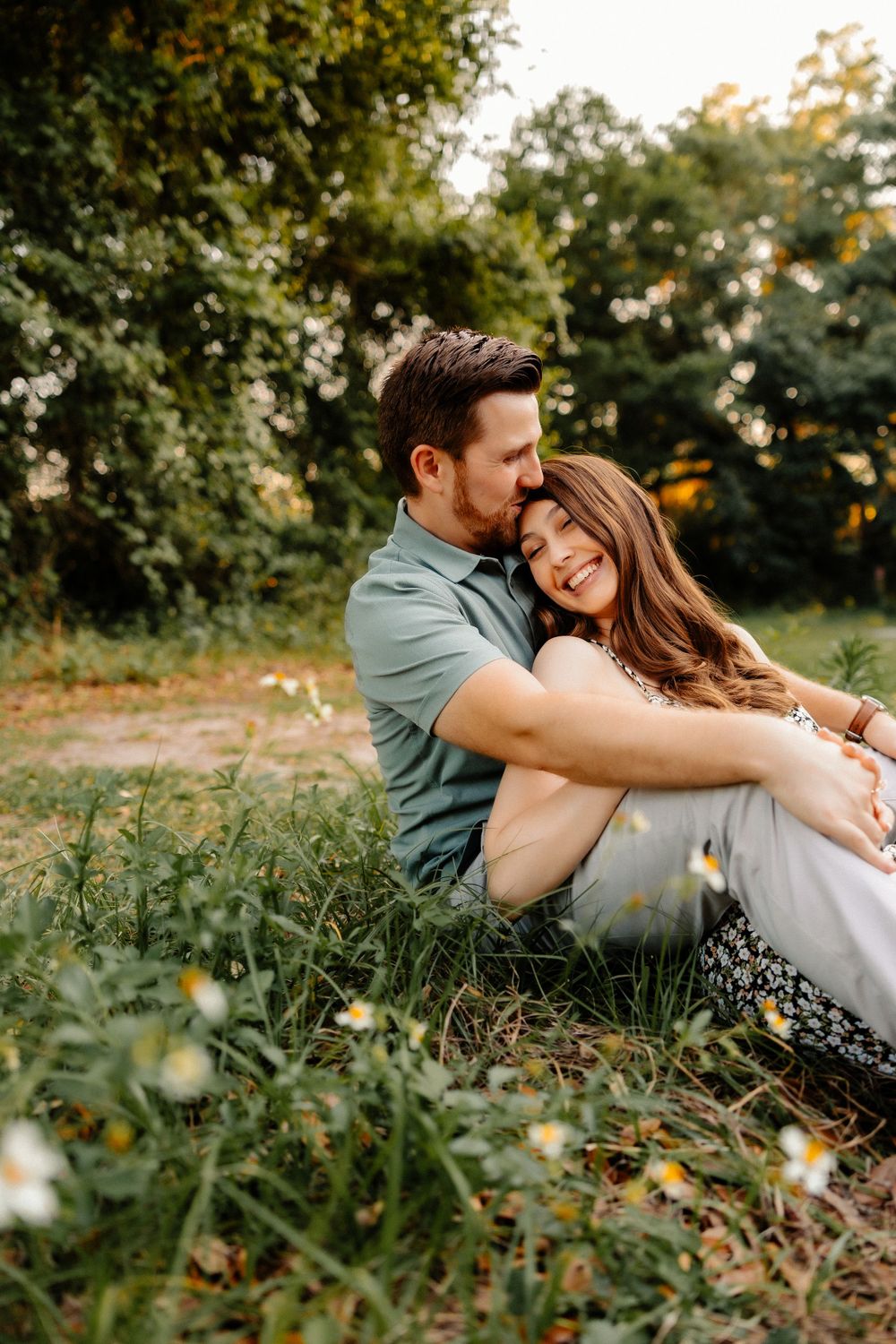 Couple sits together in a wildflower meadow, woman resting her head on her partner's shoulder.