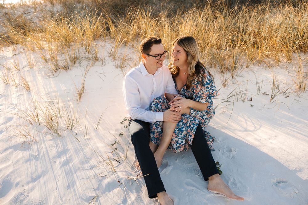 Engaged couple sitting barefoot on white sand dunes, smiling at each other during golden hour beach session.