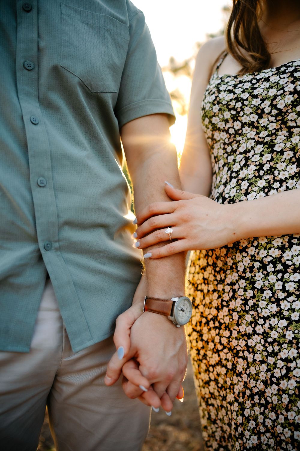 Close-up of engaged couple holding hands at golden hour, showcasing diamond engagement ring and leather watch.