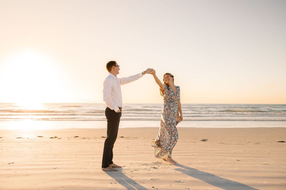 Couple dancing and twirling on the beach at sunset, woman in floral maxi dress holding bouquet.