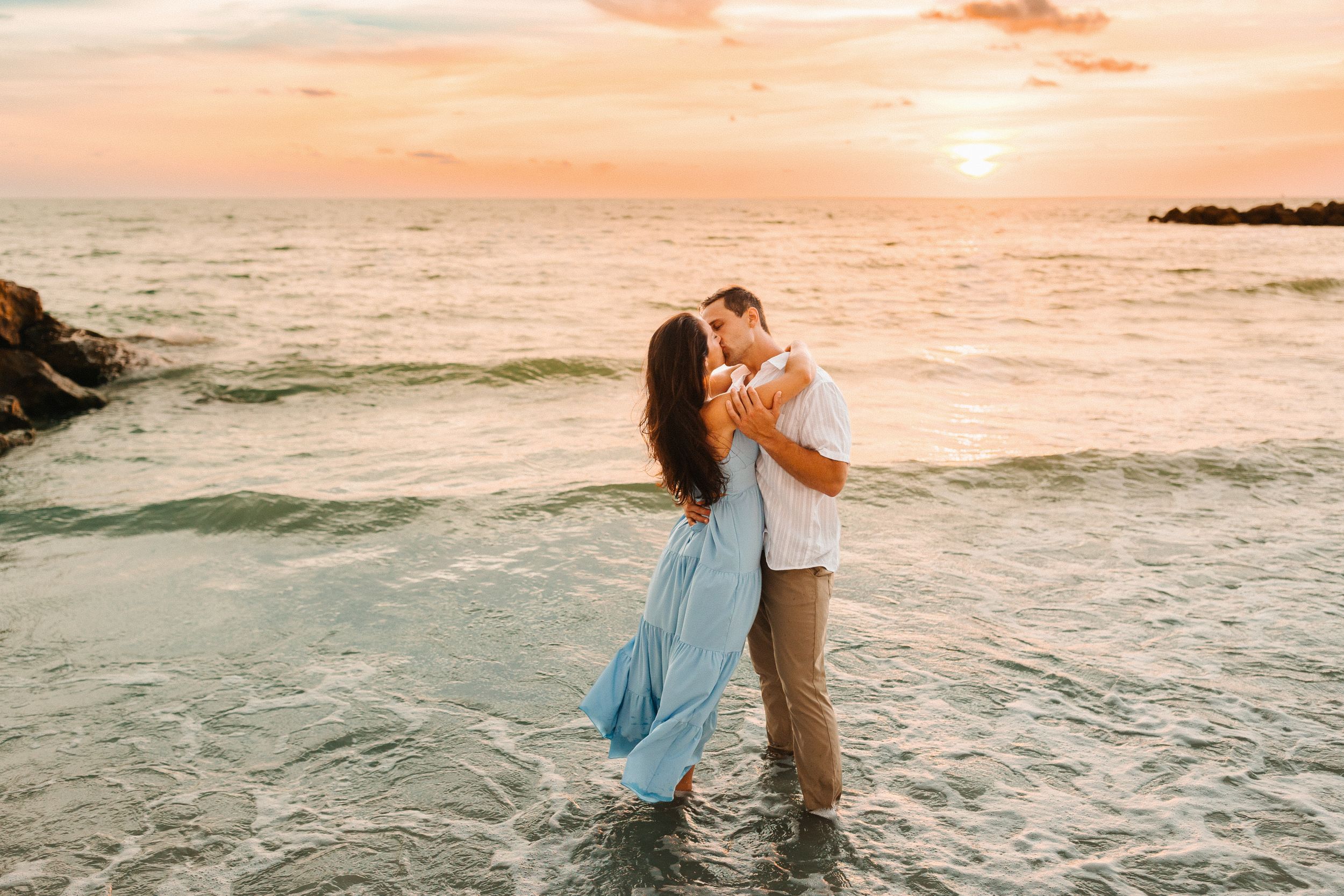 Couple kissing at sunset on a beach, standing in shallow ocean waves, woman in flowing blue dress, romantic golden sky.