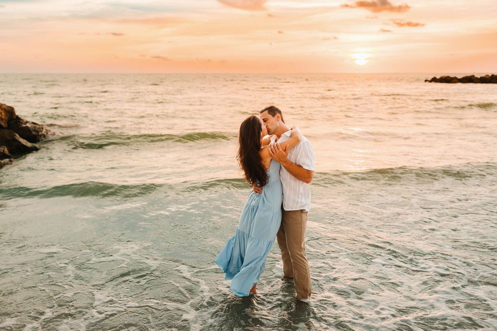 Couple kissing at sunset on a beach, standing in shallow ocean waves, woman in flowing blue dress, romantic golden sky.
