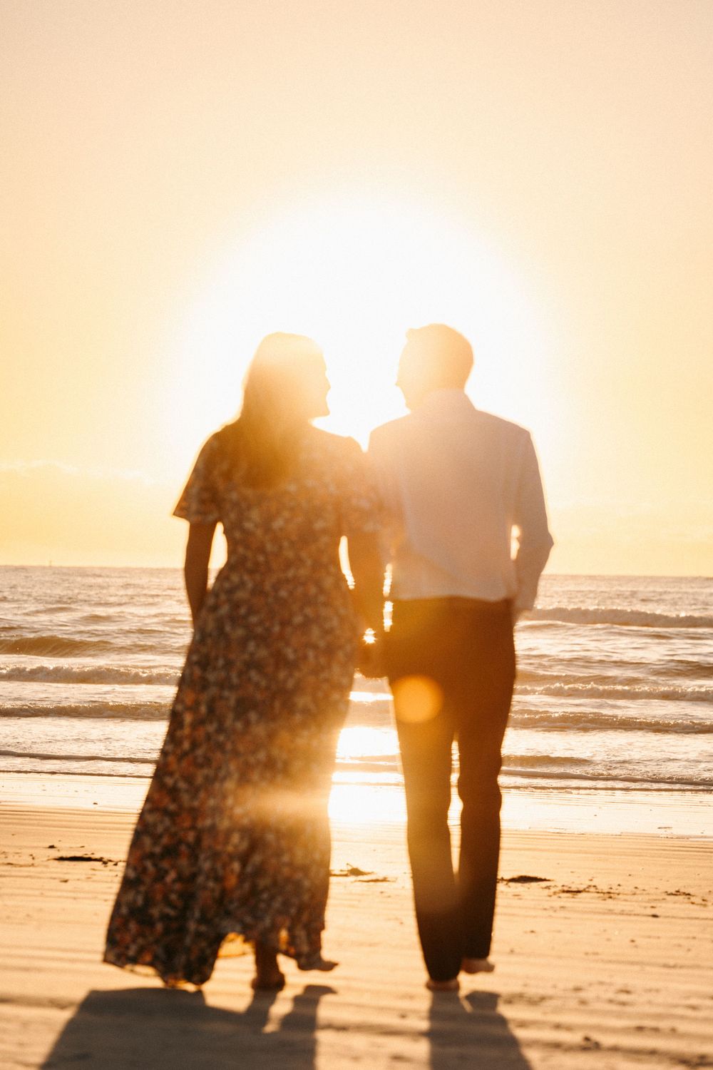 Silhouette of engaged couple holding hands walking toward the ocean during a stunning golden sunset.