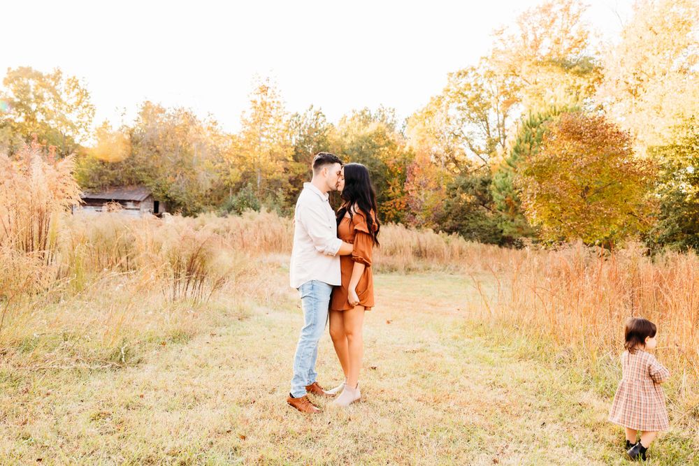 A couple embraces in a golden autumn field while a small child plays in the background during sunset.
