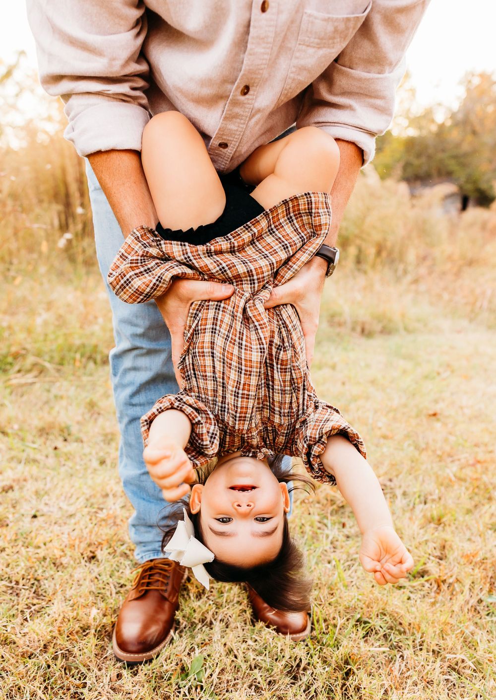 A playful moment captured in a grassy field showing someone being held upside down in a plaid shirt and jeans.