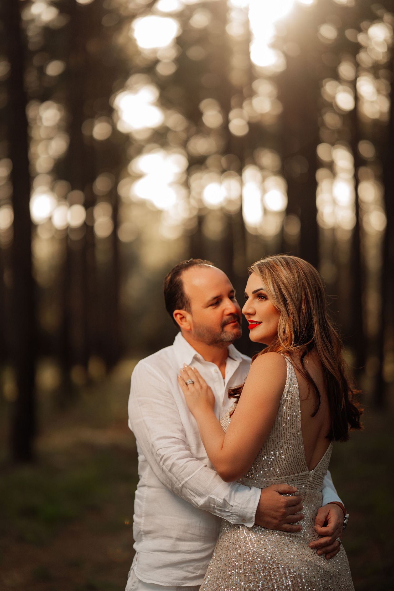 Romantic couple embraces in a dreamy forest setting during golden hour with soft bokeh lights in background.